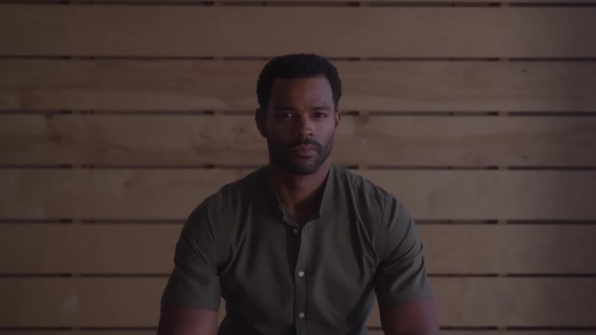 A closeup shot of a man sitting in front of a wooden wall he is looking directly at the camera with a neutral expression