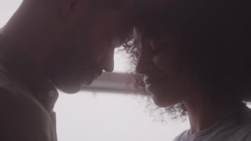 Young black couple indoors at midday standing beside a window with their foreheads gently touching smiling warmly as they share a romantic moment