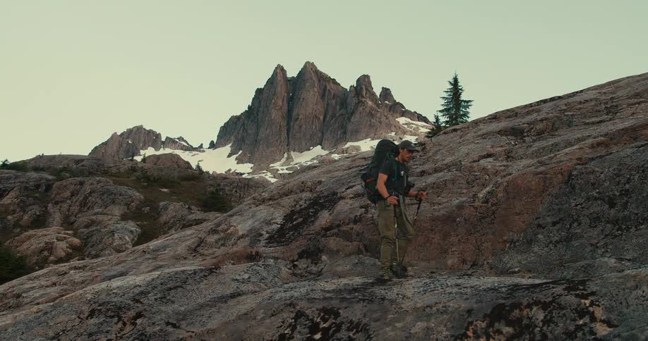 Male backpacker uses walking sticks while hiking in the mountainous terrain near a snow dusted mountain