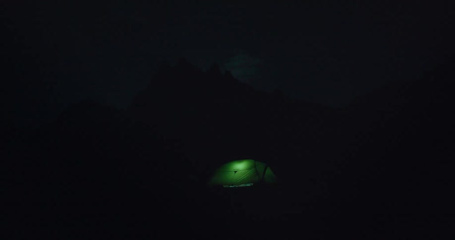 In the deep darkness of night a lone camping tent glows with a green light from within set against the faint silhouette of a mountain range