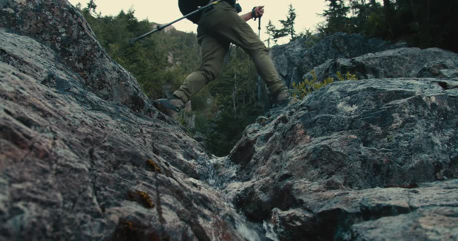 From a low angle a hiker uses trekking poles to carefully climb over wet rocks and cross a small mountain stream