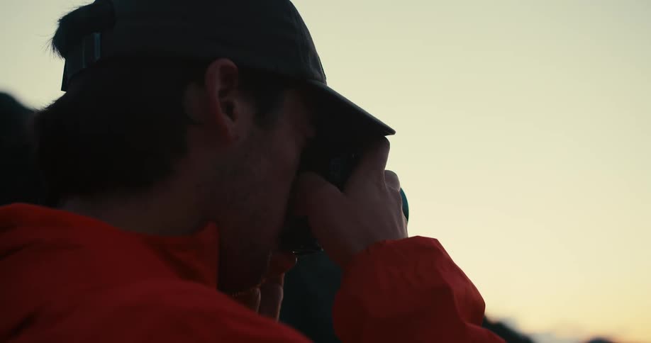 A man wearing a baseball cap and an orange jacket is looking through a camera during dusk