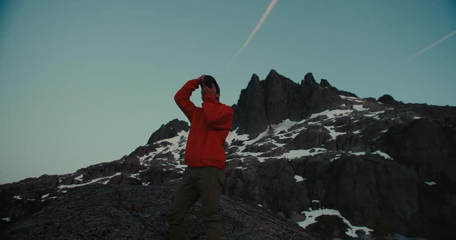 A male hiker stands on a rocky mountain holding a camera to his face and taking a photo he is wearing an orange jacket and is silhouetted against a mountain range the sky is visible behind the peaks
