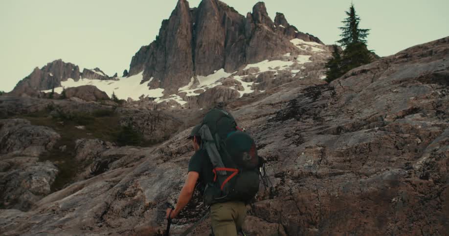Hiker with backpack treks on rocky ground toward a mountain peak