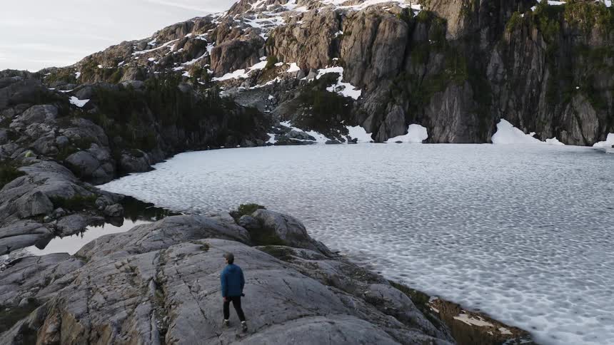 A male hiker in a blue shirt is walking along a rocky edge near a frozen lake with a large snowcovered mountain in the background