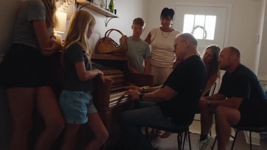 A large family gathered in a hallway around the grandfather playing piano