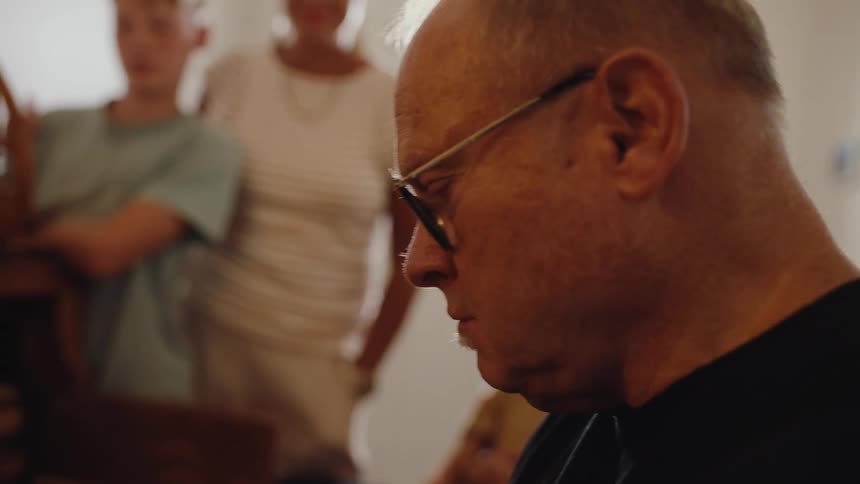 An older man wearing glasses looks down in concentration while people stand nearby watching him
