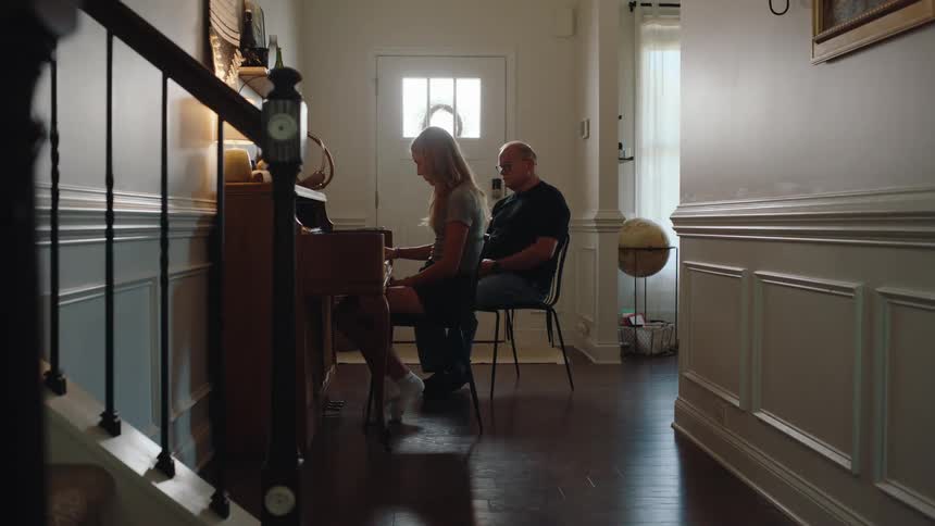 Man watches a girl playing the piano in a home room lit by natural light