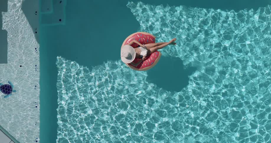 A woman wearing a sun hat is floating on a pink donutshaped inner tube reding a book in a clear blue swimming pool on a sunny day