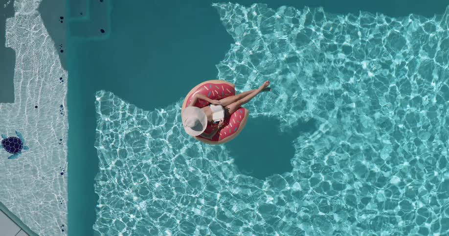 A woman wearing a sun hat is floating on a pink donutshaped inner tube in a clear blue swimming pool on a sunny day