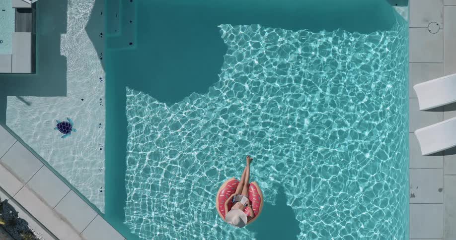 A woman wearing a sun hat is floating on a pink donutshaped inner tube reading a book in a clear blue swimming pool on a sunny day