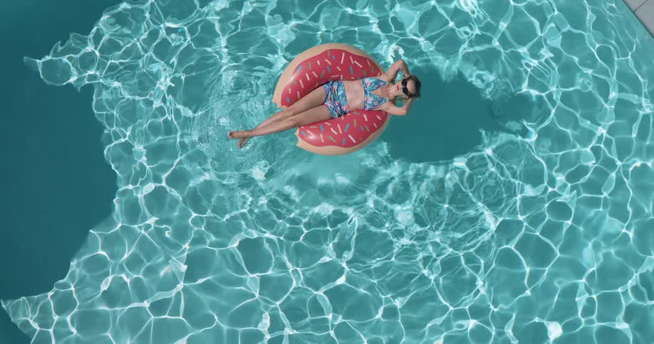 A woman in a bikini and sunglasses floats on a donut shaped inflatable in a sparkling blue swimming pool