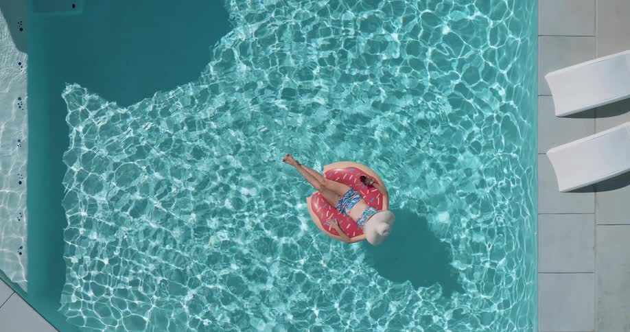 An overhead shot shows a woman wearing a sun hat and a swimsuit relaxing on a donutshaped inflatable raft in a swimming pool she is floating serenely as the sun creates ripples on the waters surface