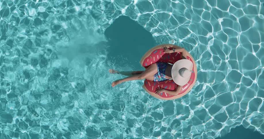 An overhead shot captures a woman on a donutshaped floatie with her legs and arms extended the woman is wearing a sun hat and a twopiece swimsuit slowly drifting across the rippling bright blue pool