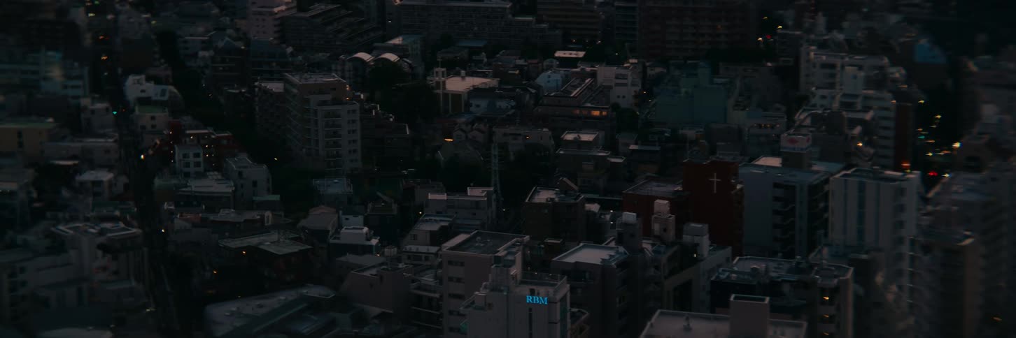 The video shows an aerial view of a city during the evening with a mix of residential and commercial buildings many of the windows are dark but some are beginning to light up indicating that the sun has set the camera is static providing a still vie