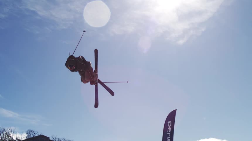 A skier is performing an acrobatic trick high in the air against a bright blue sky with the snowcovered slopes and other skiers visible in the background