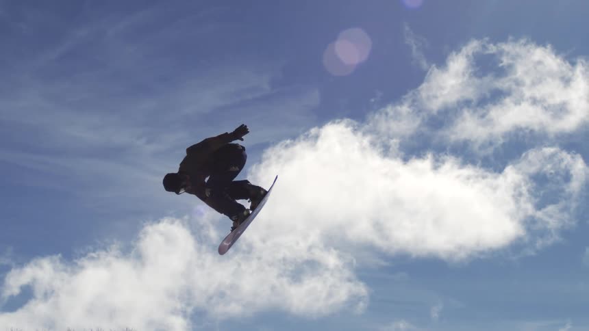 A snowboarder in dark winter clothing is performing a jump and spinning in the air against a bright blue sky with white clouds at a ski resort