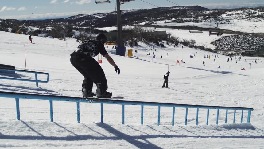 A snowboarder is sliding down a blue handrail on a snowy slope with ski lifts and people on the mountain visible in the background on a sunny day