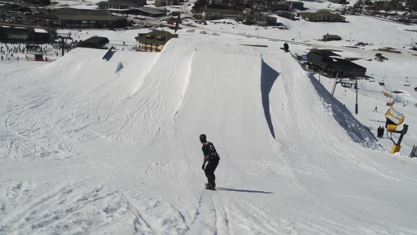 A snowboarder in dark winter clothing is riding down a snowy slope toward a large jump at a ski resort