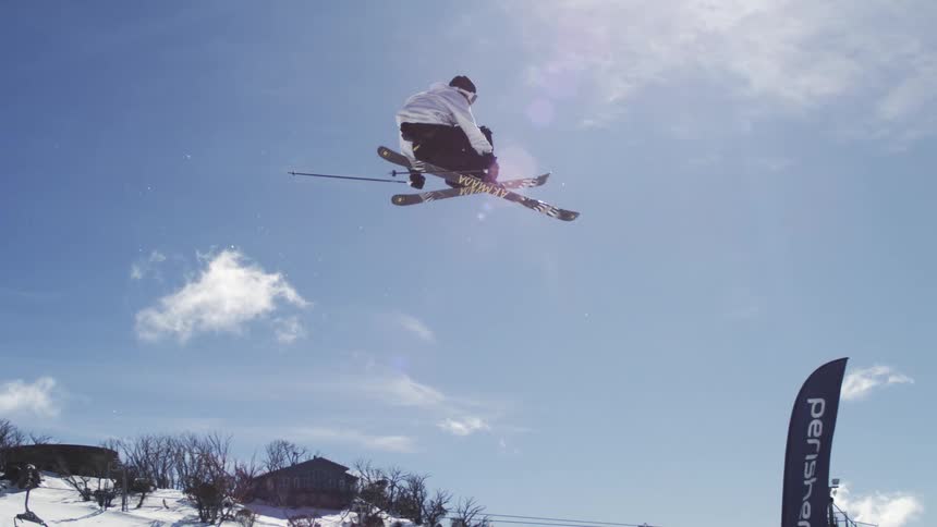 A skier in white pants and a black jacket is performing an acrobatic trick and spinning in the air with a ski resort and mountains visible below him
