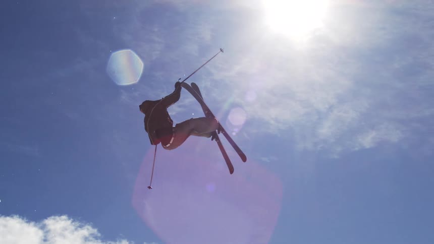 In the first shot a skier executes a midair backflip against a bright blue sky with a ski resort visible on the snowy ground below the second shot is a closeup of the skier now descending their silhouette sharp against the sun as they hold