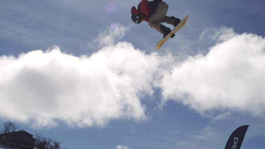 Snowboarder launching off the snowy slope into the air soaring high above the snowfilled mountains at midday under a bright sunny sky