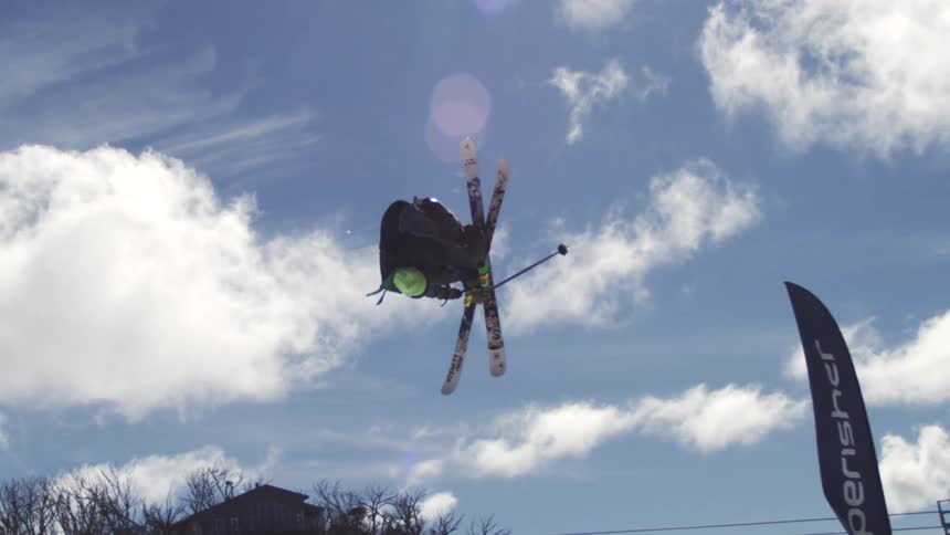 A skier performs an aerial trick against a blue sky with clouds landing in a snow covered landscape with ski lifts and buildings in the background