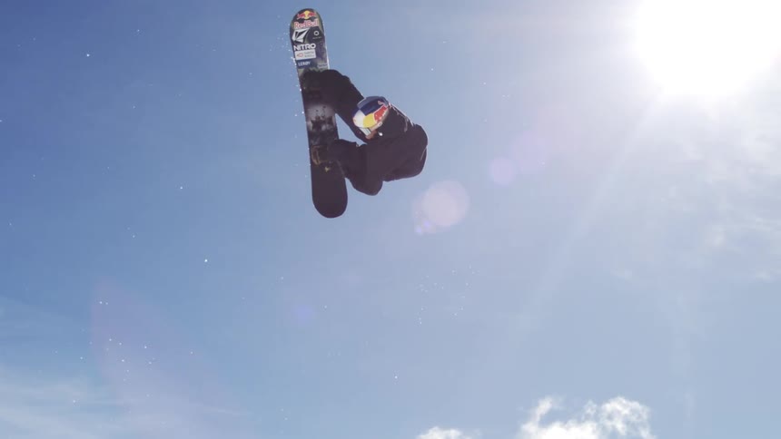 A snowboarder is performing an acrobatic trick high in the air against a bright blue sky with the snowcovered slopes and a building visible in the background