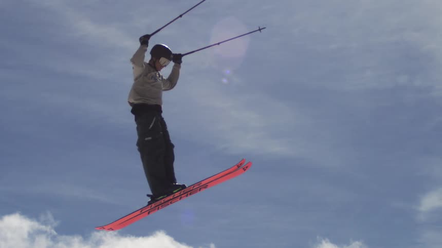 Man wearing a helmet and ski gear flung into the air while holding two ski poles midjump on a sunny winter day in the snowy mountains