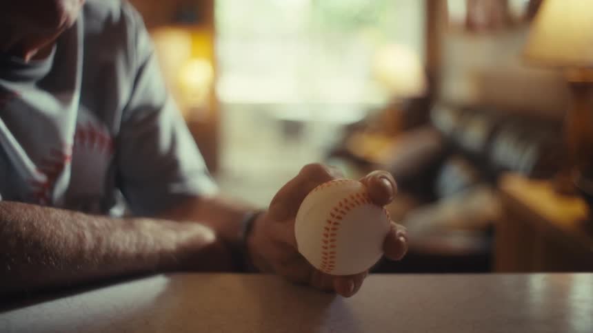 A close view of a mans hand shows him holding a baseball at a table with a calm and nostalgic atmosphere with the interior of his home in the background