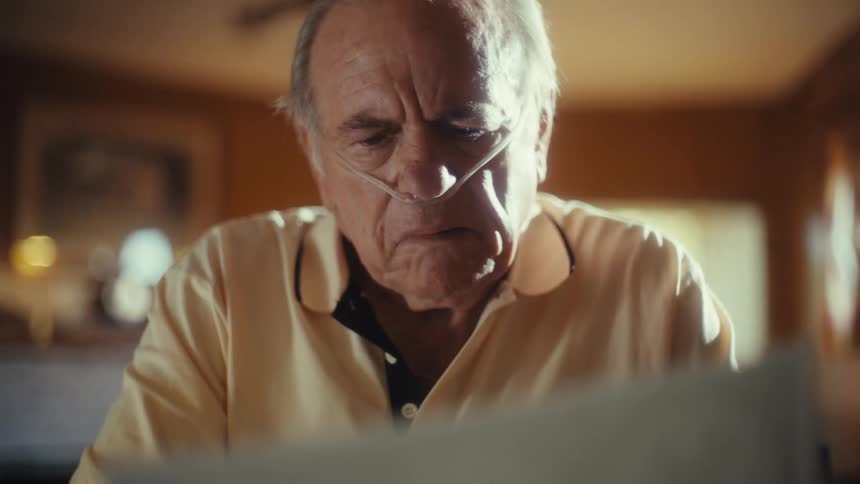 A sick elderly man with a nasal cannula looks up from a medical brochure at someone offscreen he then turns his gaze back to the brochure which is for fsp health