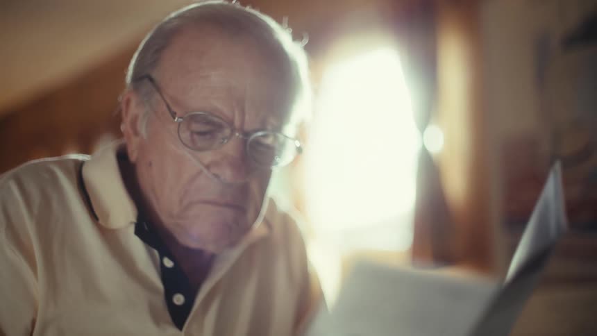 An elderly man wearing glasses and a nasal cannula is sitting in his home and reading a brochure about comprehensive care