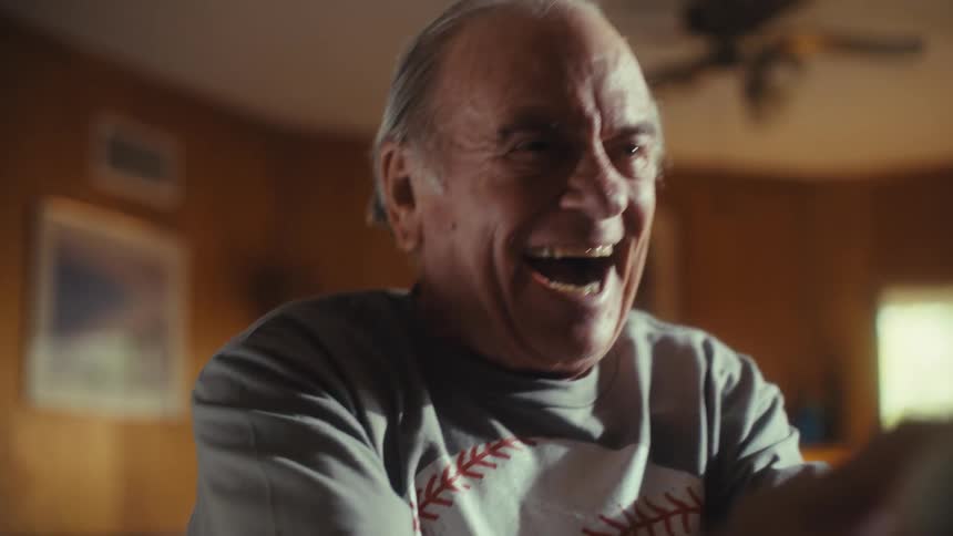 A man in a baseball shirt sits at a counter watching something offscreen he throws his head back and laughs loudly in a moment of genuine joy and excitement