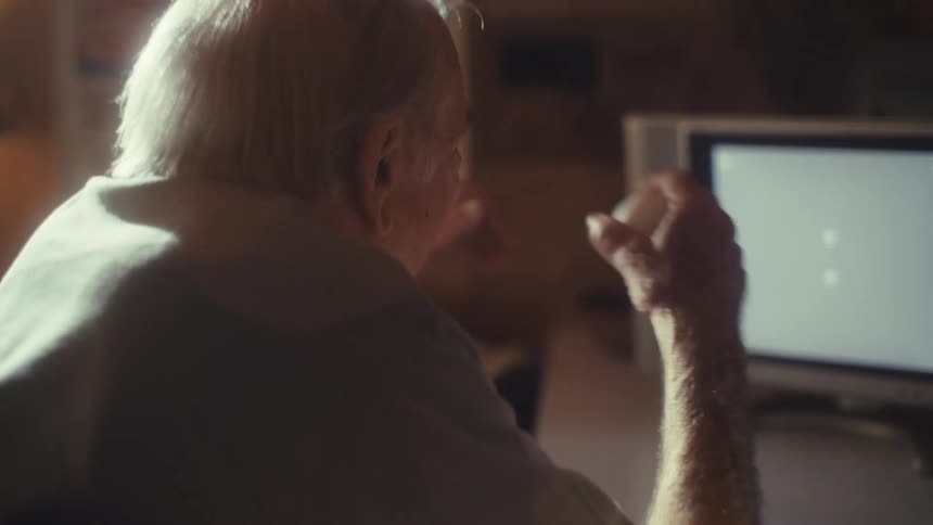 An elderly man gestures excitedly toward a computer screen in a warmly lit room