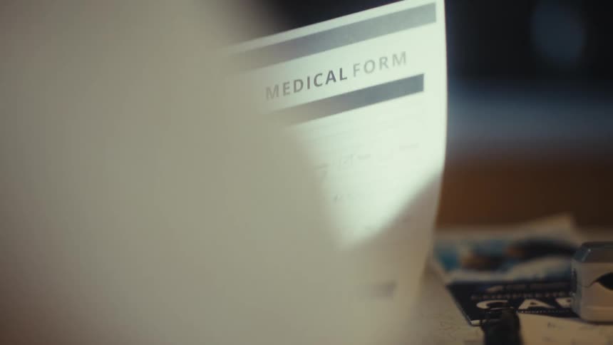 A blurry over the shoulder view shows a persons hand filling out a medical form with a pen on a wooden table