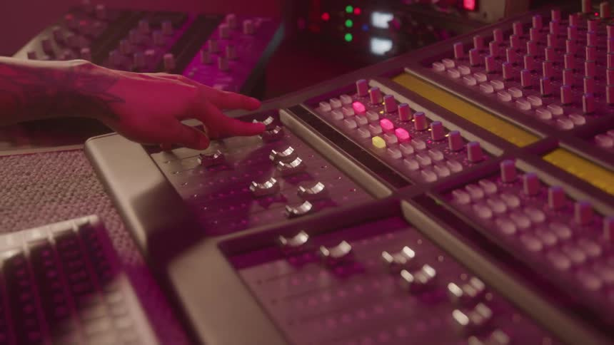 A persons tattooed hand adjusts controls on an illuminated audio mixing console