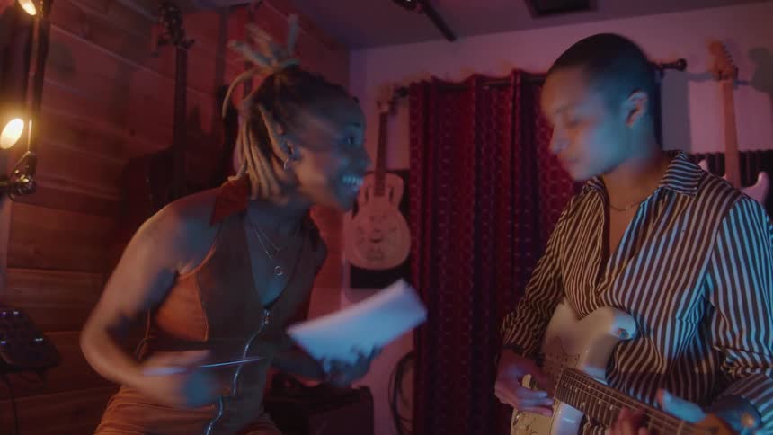 Two women interact in a music studio while one holds notes and the other plays guitar