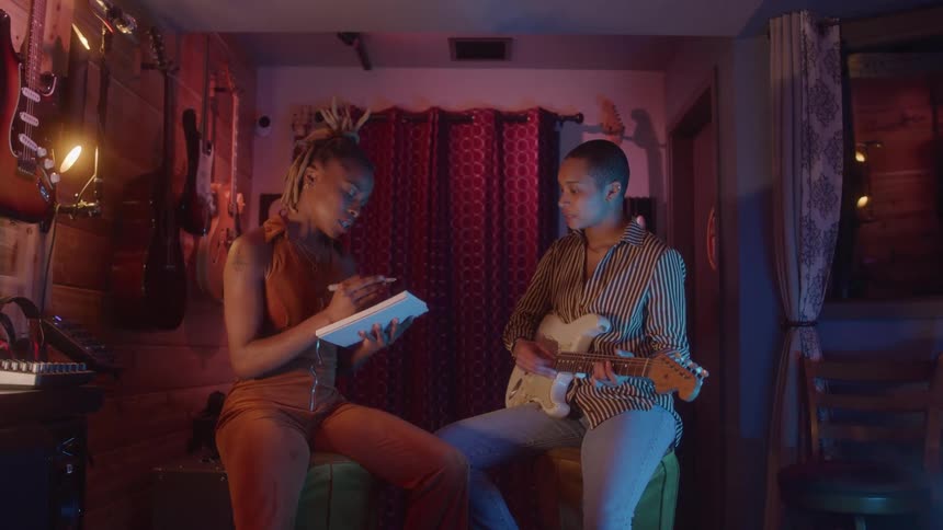 Two women are sitting and collaborating on a song in a music studio with one woman playing an electric guitar and the other holding a notebook and a pen