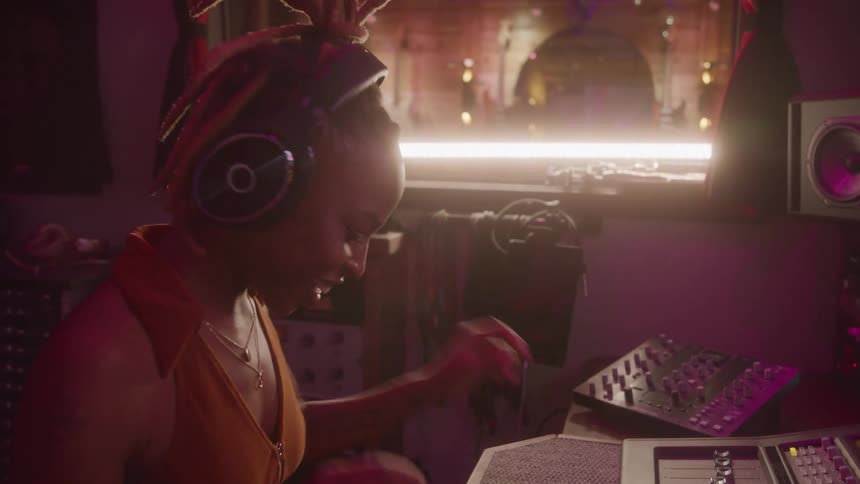 A woman with dreadlocks and headphones is sitting at a mixing board in a dimly lit music studio listening to music and bobbing her head