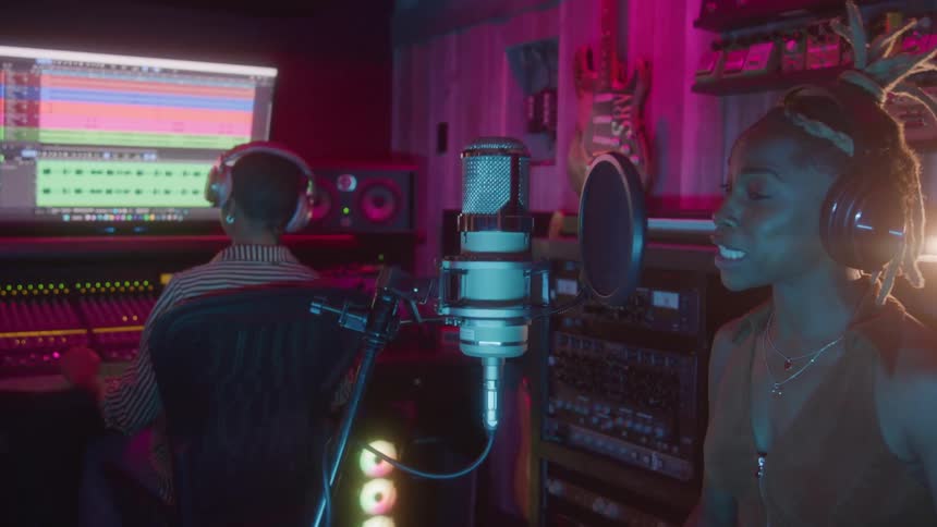 A woman wearing headphones is smiling and singing into a studio microphone while a man at a mixing console works on audio production in the background