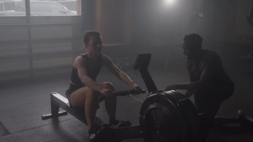 A woman is working out on a rowing machine in a dark gym with a man next to her coaching her as the sunlight from a large window creates a hazy and dramatic atmosphere