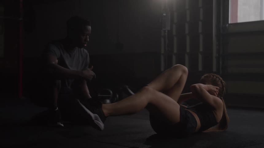 A female athlete in black sportswear is doing weighted situps on an exercise mat in a dark gym with a male coach holding her feet and giving her a red medicine ball