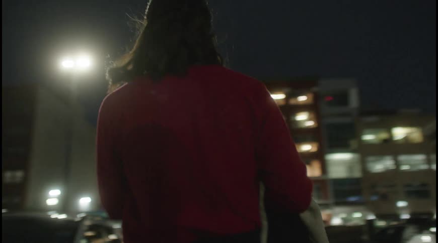A woman walks through a parking lot at night with cars and buildings behind