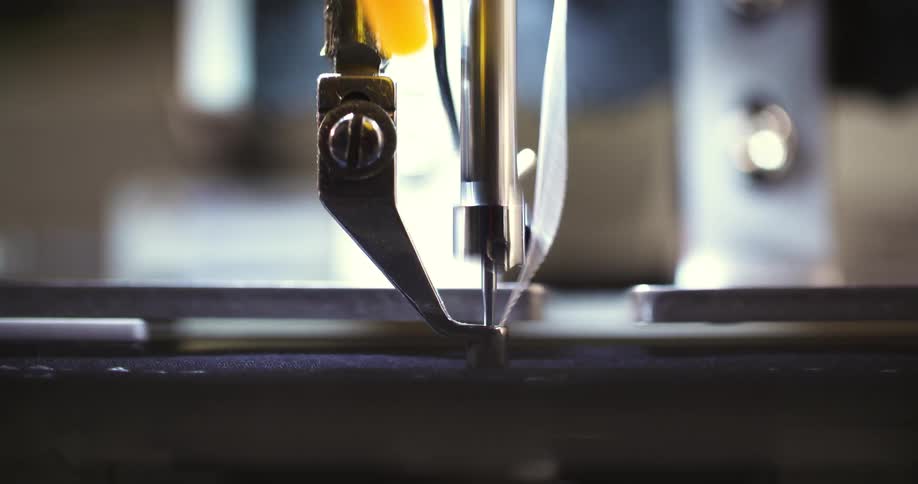 A sewing machine needle moves up and down while a thread creates a stitch on a piece of dark fabric