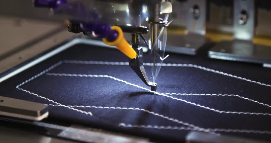 A computerized sewing machine stitches precise white zigzag patterns onto dark fabric with mechanical accuracy