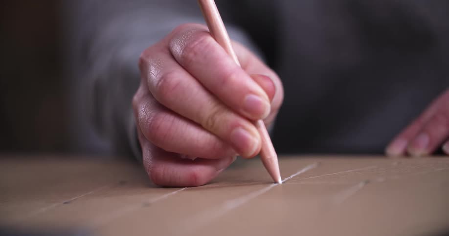 A person is using a pinkcolored pencil to draw a straight line on a piece of cardboard the camera is focused on their hand and the pencil as it moves across the surface
