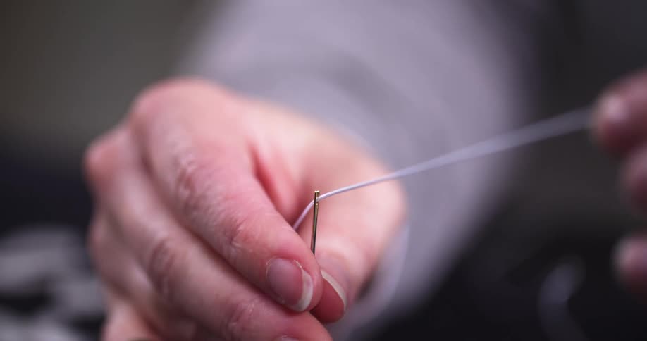 A close view shows a persons hands pulling a thread through the eye of a needle