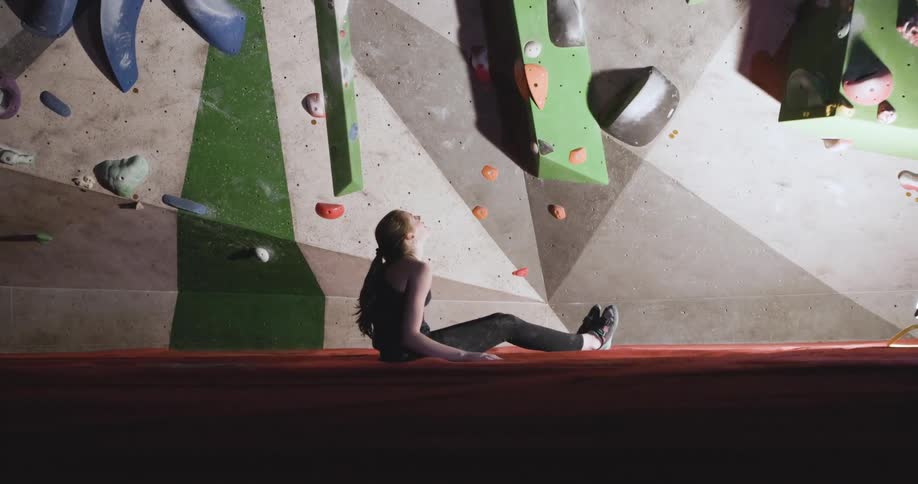 A young girl sitting and looking to a rock climbing wall in a indoor setting