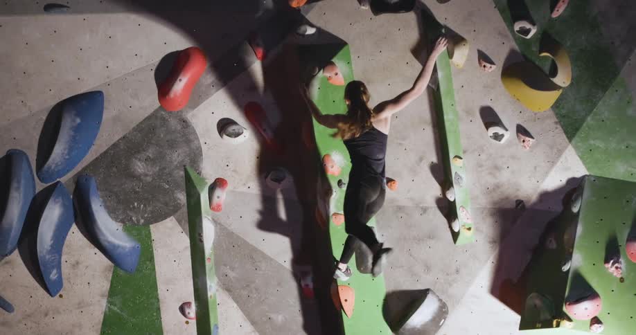 Young woman with long blonde hair wearing black pants and a black singlet doing indoor rock climbing swerving from one handhold to another on the wall