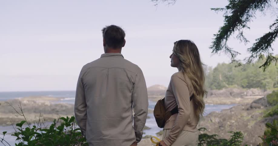 Couple standing on a beach shoreline during vacation looking at the distant ocean view at midday as a light wind blows through the womans long blonde hair